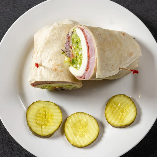 A Vinnie's Pizzeria wrap, cut in half, revealing meat, cheese, and lettuce, served with pickle slices.