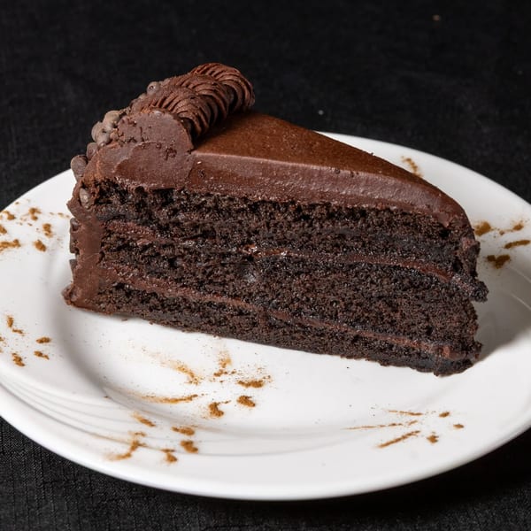 A slice of rich chocolate cake with chocolate frosting and decorative piping on a white plate.