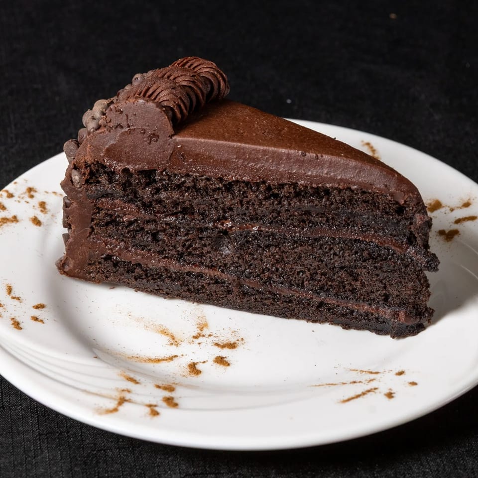A slice of rich chocolate cake with chocolate frosting and decorative piping on a white plate.
