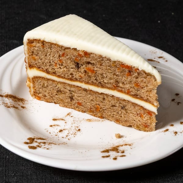A slice of carrot cake with white frosting and cinnamon dusting on a white plate.