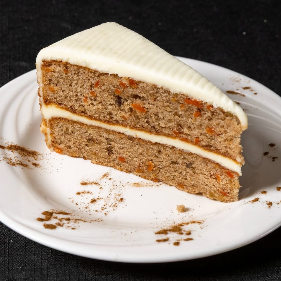 A slice of carrot cake with white frosting and cinnamon dusting on a white plate.
